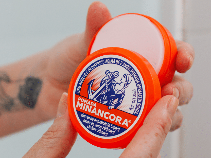 Mãos segurando o potinho laranja da Pomada Minancora com o novo logotipo. 