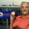 Foto da Dona Elma, usando uma camisa laranja, segurando uma pomada Minancora com o grafismo "110 anos Minancora" ao lado