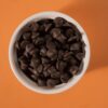 Porção de gotas de chocolate em tigela branca sobre fundo laranja, ilustrando a relação entre consumo de chocolate e o tema 'chocolate causa espinhas'.