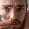 Homem com barba espessa segura um barbeador próximo ao rosto, com expressão concentrada. A lâmina está posicionada perto da bochecha, sugerindo o momento antes ou durante o barbear.