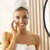 Mulher sorridente em ambiente de spa ou banheiro, com toalha branca no corpo e na cabeça, usando patches para os olhos, representando uma rotina de skincare 30 anos.