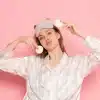 Mulher com pijama de cetim claro e máscara de dormir com orelhas, segurando discos de algodão em frente a fundo rosa, representando o ritual noturno do sono da beleza.