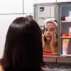 Pessoa aplicando produto facial diante do espelho, com faixa rosa na cabeça, representando uma rotina de skincare minimalista em ambiente de banheiro.